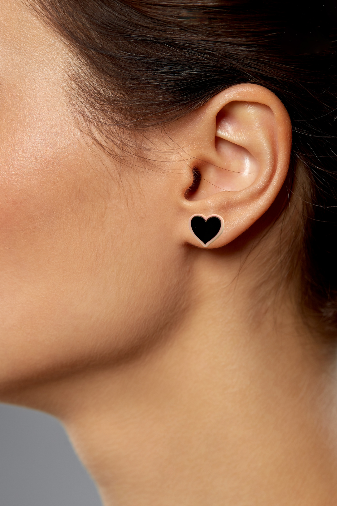 A pair of pastel pink heart shaped acrylic earrings with a bold black heart being worn in the ear lobe of a woman facing to the side.