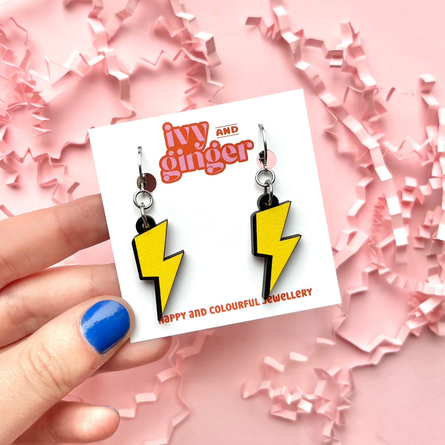 A photograph of two lightning bolt shaped dangle earrings connected to a small huggie hoop presented on a white earring card with the brand name 'ivy and ginger' at the top being held by someone.