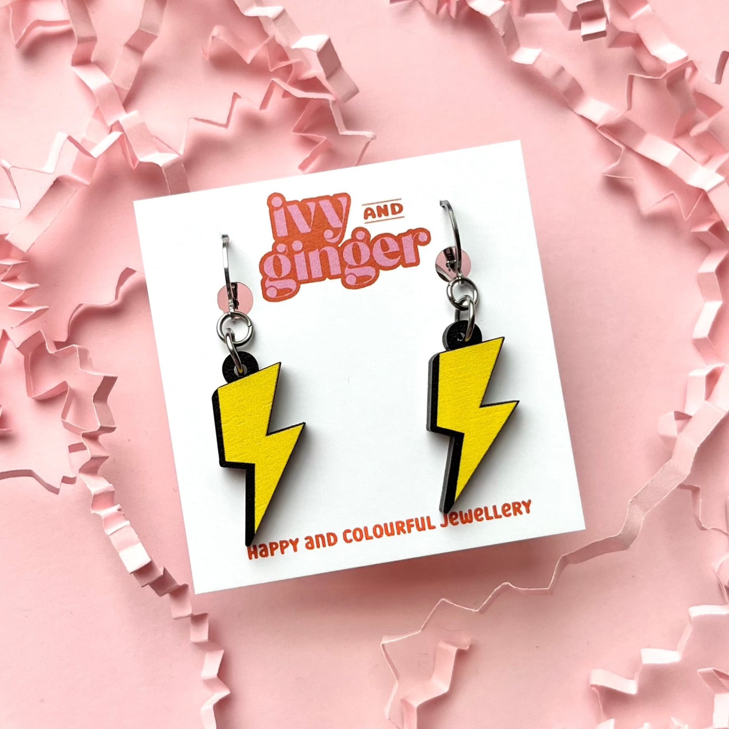 A photograph of two lightning bolt shaped dangle earrings connected to a small huggie hoop presented on a white earring card with the brand name 'ivy and ginger' at the top on a pink background.