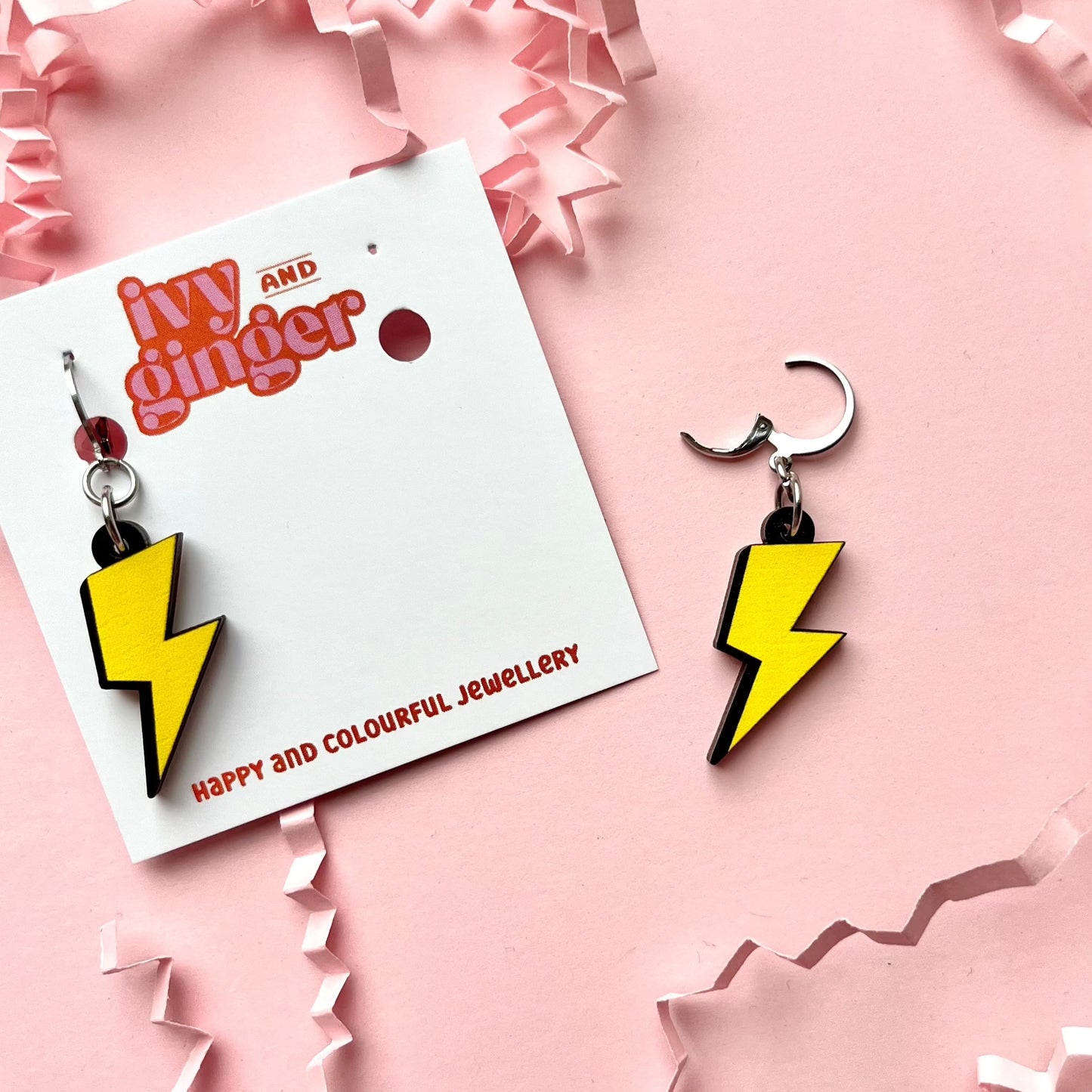 A photograph of two lightning bolt shaped dangle earrings connected to a small huggie hoop. One earring is attached to a white earring card with the brand name 'ivy and ginger' at the top. The other is lying next to the card on a pink background.