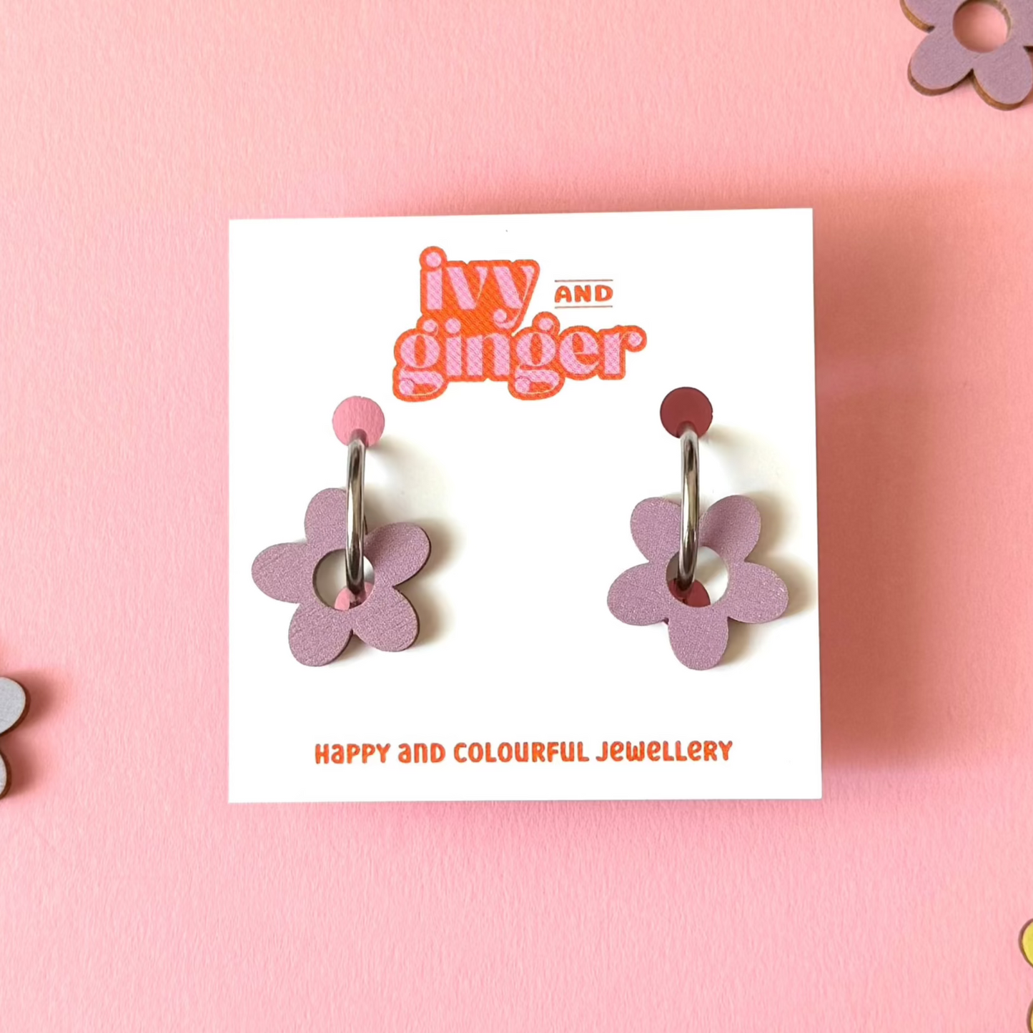 Dusky lilac coloured wooden flower shapes attached to a steel hoop presented on a white backing card with the brand name 'ivy and ginger' at the top. The earrings are placed on a pink background with extra wooden flower shapes placed around the photo.