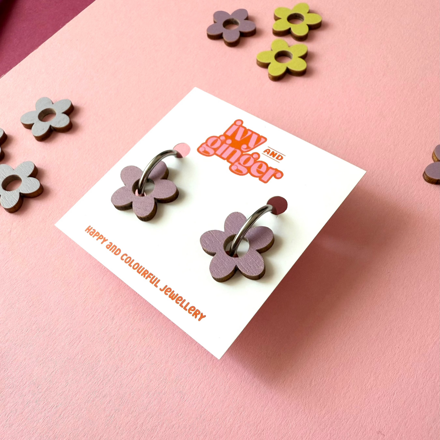 Dusky lilac coloured wooden flower shapes attached to a steel hoop presented on a white backing card with the brand name 'ivy and ginger' at the top. The earrings have been placed at an angle so we can see them close up. The earrings are placed on a pink background with extra wooden flower shapes placed around the photo.