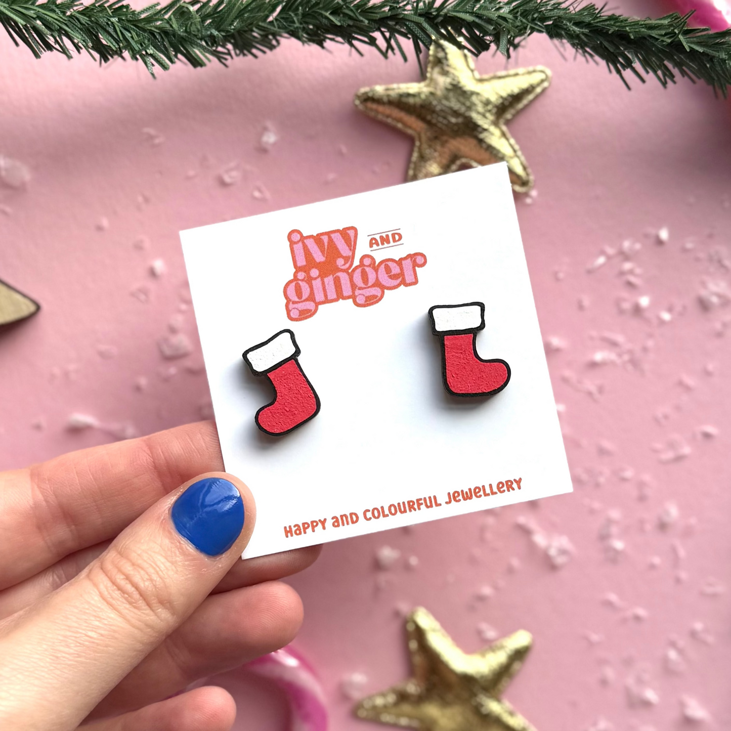 Card with red and white stocking earrings held by a hand on a pink background with Christmas decorations.