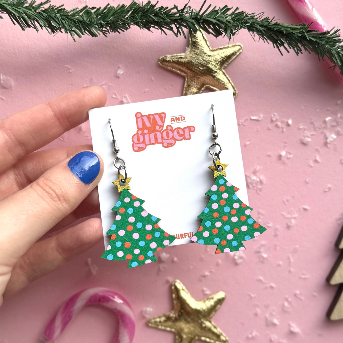 Hand holding a card with Christmas tree earrings on a pink background with festive decorations.