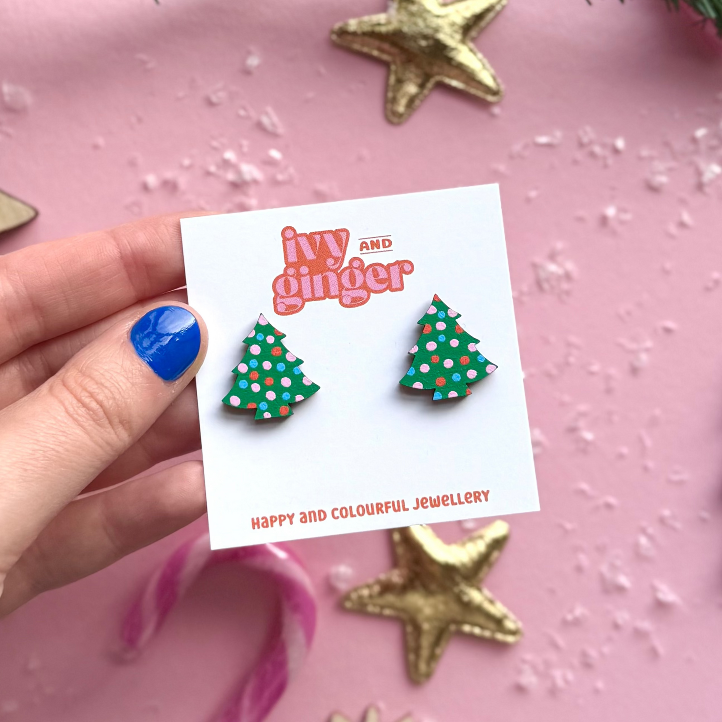 Hand holding a card with Christmas tree earrings and 'Ivy and Ginger' branding on a pink background.