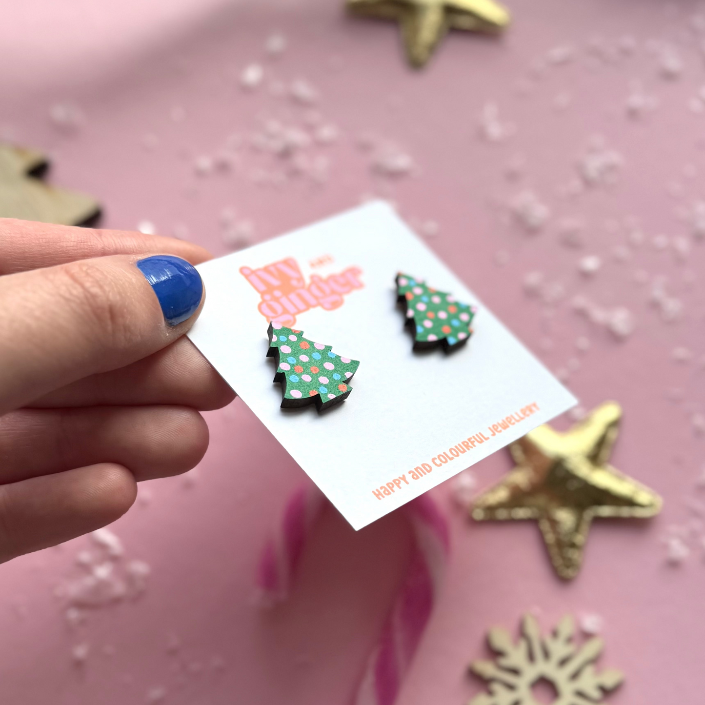 Hand holding a card with Christmas tree earrings on a pink background with star decorations.