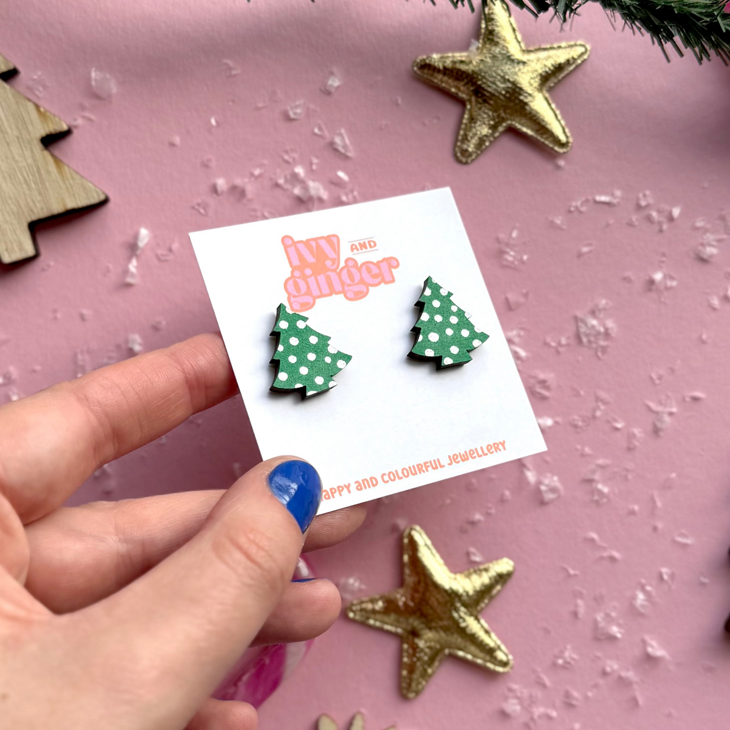 Hand holding a card with green polka dot earrings on a pink surface with Christmas decorations.