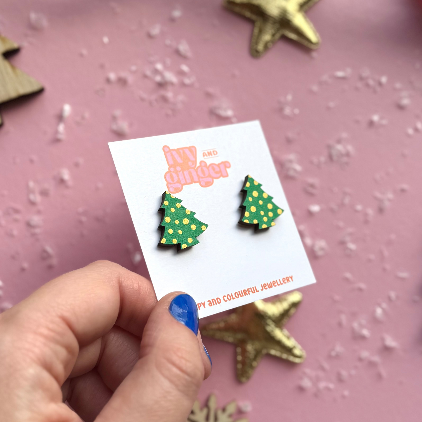 Hand holding a card with Christmas tree earrings against a pink background with decorative elements.