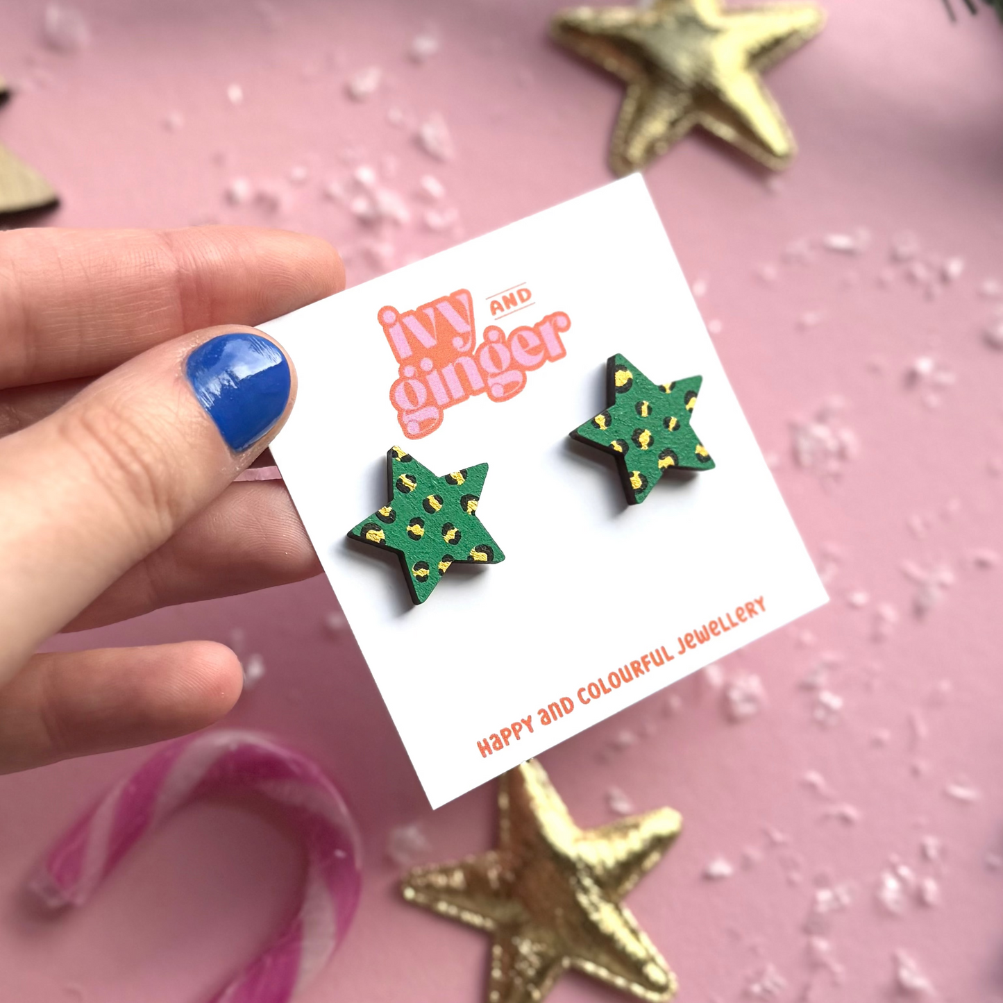 Hand holding a card with green star-shaped earrings on a pink background with gold stars.
