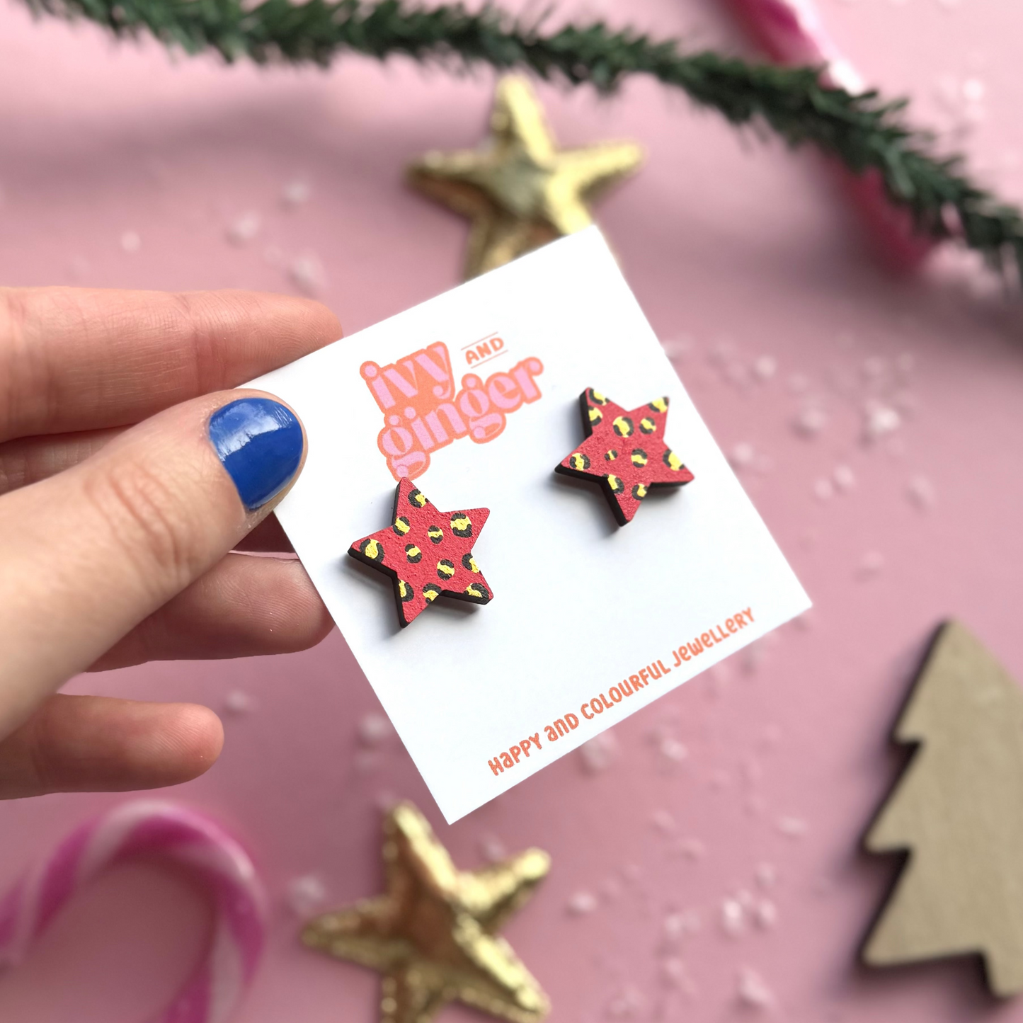 Star-shaped earrings on a 'Jewellery' card held by a hand with blue nail polish, against a pink background with decorative elements.