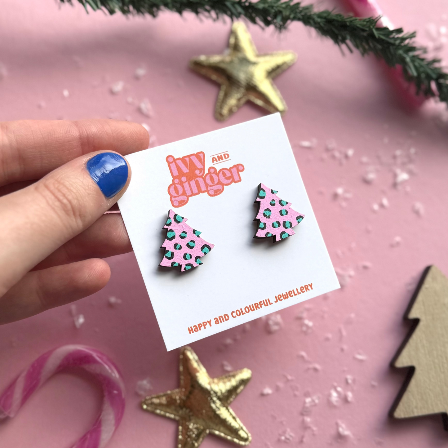 Hand holding a card with Christmas-themed earrings on a pink background with decorative elements.