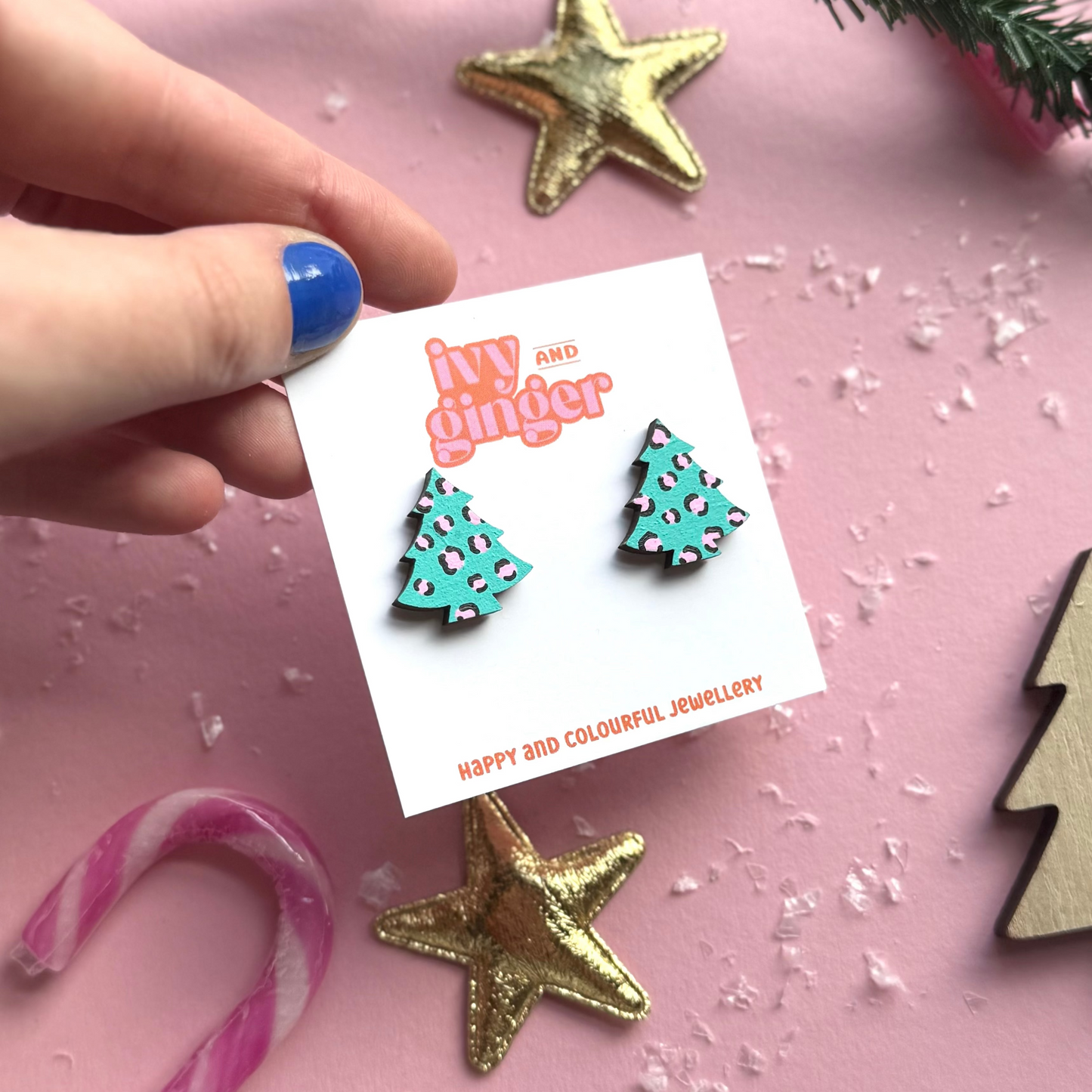 Turquoise Christmas tree earrings on a card with 'iw and ginger' branding, surrounded by festive decorations on a pink background.