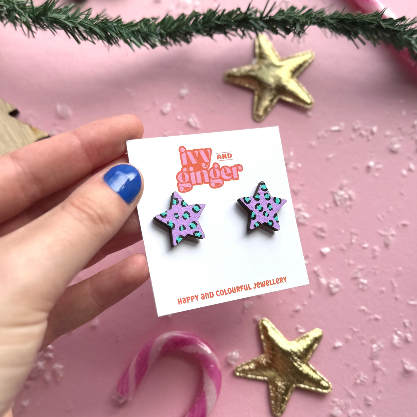 Star-shaped earrings on a card held by a hand with blue nail polish, against a pink background with decorative elements.