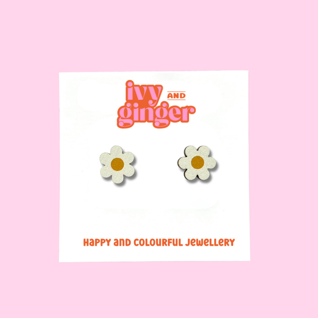 A pair of white daisy shaped wooden stud earrings with a yellow circle centre. The earrings are being displayed on a white backing card with the brand name 'ivy and ginger' at the top.
