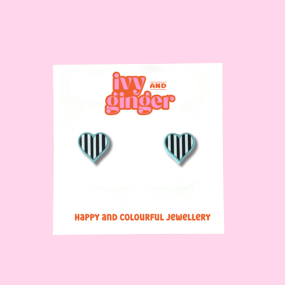 Midi-sized pastel blue heart stud earrings with black and white striped details displayed on a white backing card with the brand name 'ivy and ginger' at the top.