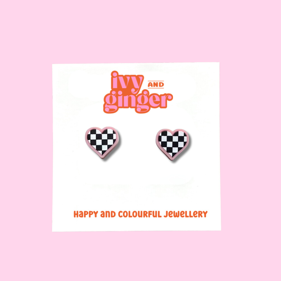 A pair of earrings featuring a bold black and white checkerboard heart design set against a smooth pastel pink acrylic base. The earrings are displayed on a white backing card with the brand name 'ivy and ginger' at the top.