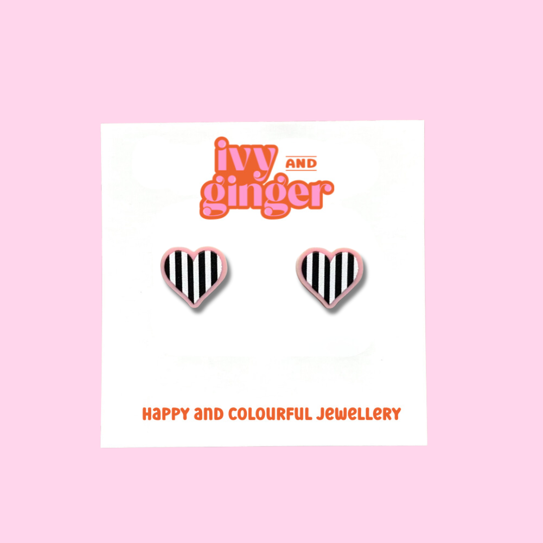 A pair of black and white striped hearts on pink acrylic heart shaped stud earrings. The earrings are displayed on a white backing card with the brand name 'ivy and ginger' at the top.