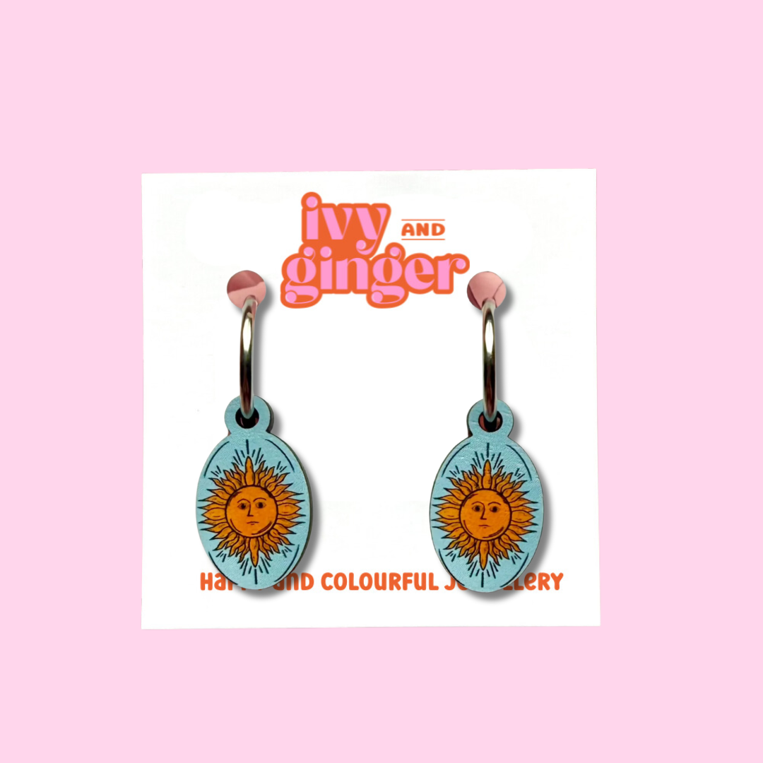 A photograph of light blue oval shaped earrings with a celestial inspired sun design attached to steel hoops, presented on a white backing card with the brand 'ivy and ginger' at the top.