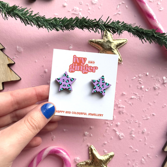 Star-shaped earrings on a card held by a hand with Christmas decorations on a pink background
