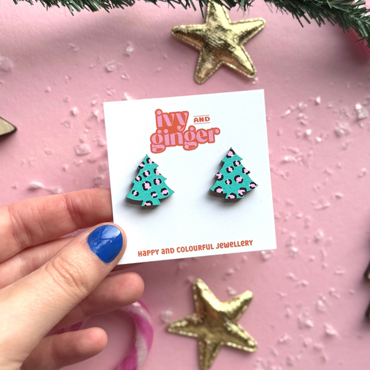Hand holding a card with Christmas-themed earrings on a pink background with decorative elements.