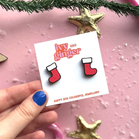 Card with red and white stocking earrings held by a hand on a pink background with Christmas decorations.