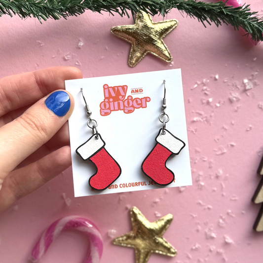 Hand holding a card with red stocking earrings on a pink background with Christmas decorations.
