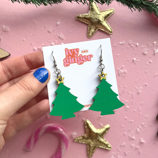 Hand holding green Christmas tree earrings with gold stars on a pink background with festive decorations.
