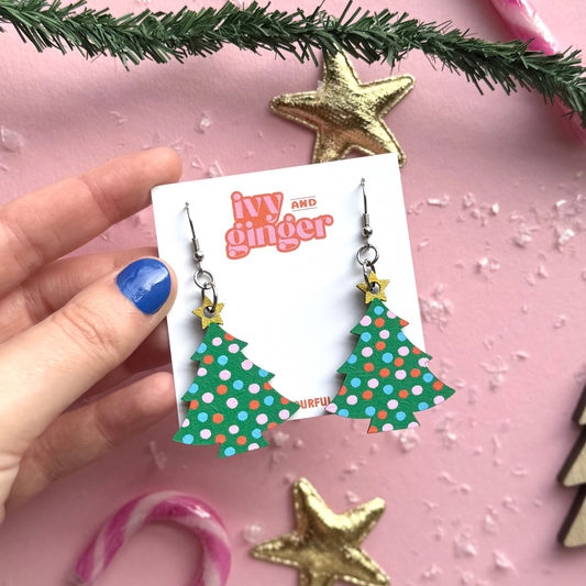 Hand holding a card with Christmas tree earrings on a pink background with festive decorations.