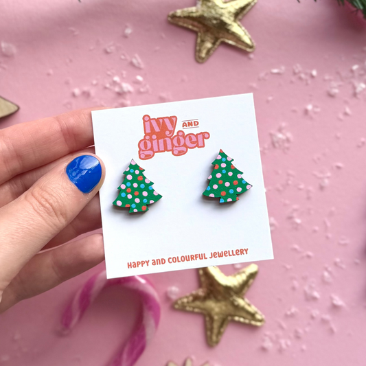 Hand holding a card with Christmas tree earrings and 'Ivy and Ginger' branding on a pink background.