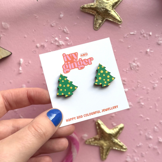 Hand holding a card with green Christmas tree earrings on a pink background with decorative elements.