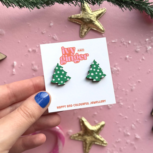 Hand holding a card with green polka dot Christmas tree earrings on a pink background with decorative elements.
